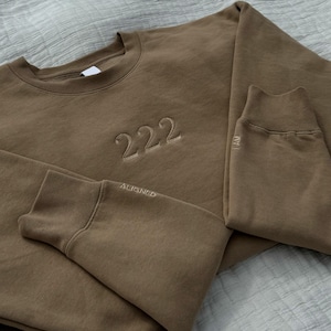 K&ouml;nnte beinhalten: Ein braunes Sweatshirt mit der Zahl "222" auf der Vorderseite gestickt. Die Worte "I AM" und "ALIGNED" sind auf den &Auml;rmeln gestickt.
