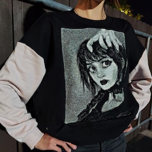 May include: Black sweatshirt with a white and black graphic print of a woman's face with a hand on her head. The sweatshirt has a white long sleeve on the left side and a black long sleeve on the right side.