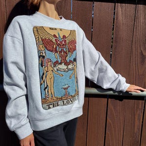 Puede incluir: Una sudadera gris con una gran imagen bordada de una carta del tarot, Los Enamorados, que presenta dos figuras y una figura alada sobre ellas. La carta est&aacute; rodeada por un borde azul y el texto "LOS ENAMORADOS" est&aacute; impreso debajo de la imagen.