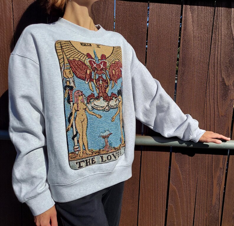 Puede incluir: Una sudadera gris con una gran imagen bordada de una carta del tarot, Los Enamorados, que presenta dos figuras y una figura alada sobre ellas. La carta est&aacute; rodeada por un borde azul y el texto "LOS ENAMORADOS" est&aacute; impreso debajo de la imagen.