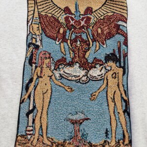 Puede incluir: Un tapiz que representa la carta de Los Enamorados de una baraja de tarot. La carta presenta dos figuras desnudas de pie a cada lado de un &aacute;rbol con una corona en la cima. Las figuras se toman de la mano y se miran. La carta est&aacute; rodeada de un marco dorado con las palabras "THE LOVERS" en la parte inferior.