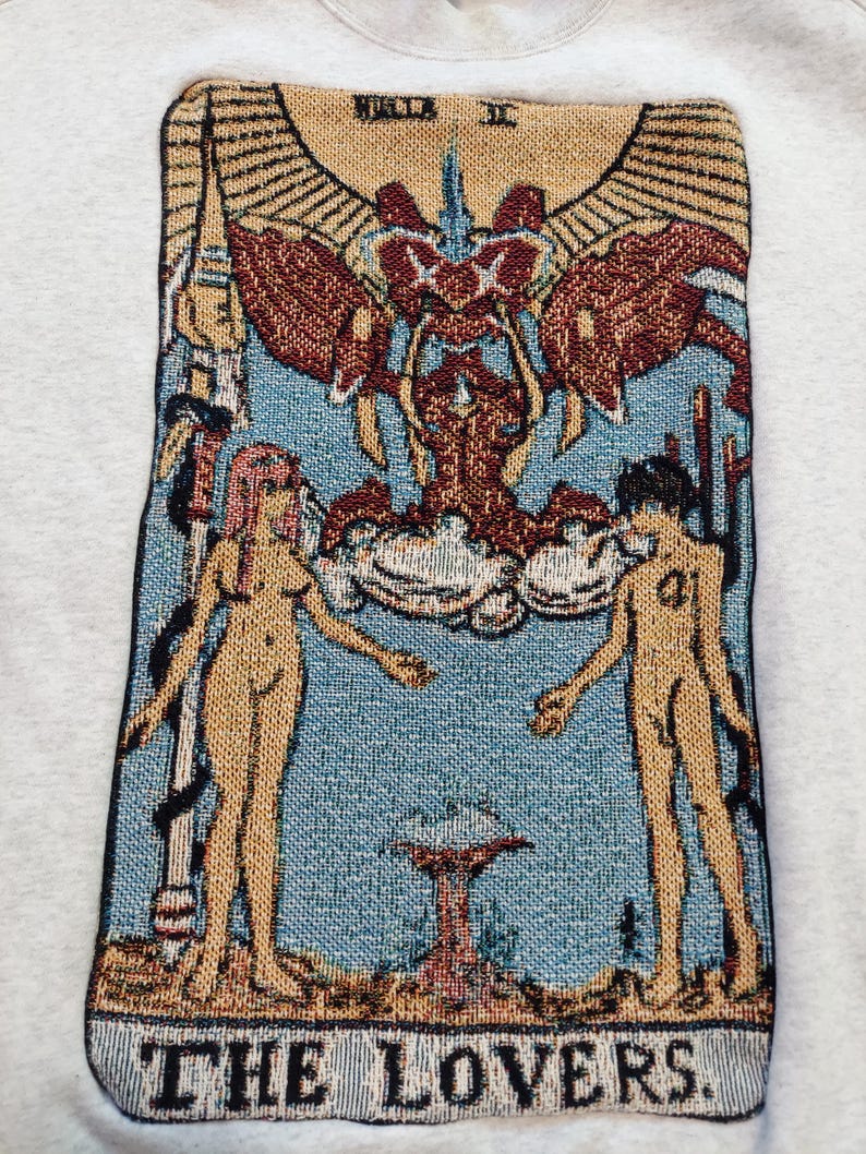 Puede incluir: Un tapiz que representa la carta de Los Enamorados de una baraja de tarot. La carta presenta dos figuras desnudas de pie a cada lado de un &aacute;rbol con una corona en la cima. Las figuras se toman de la mano y se miran. La carta est&aacute; rodeada de un marco dorado con las palabras "THE LOVERS" en la parte inferior.