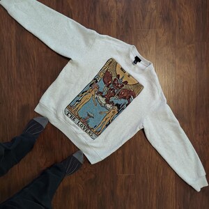 Puede incluir: Una sudadera gris claro con una gran imagen bordada de la carta del tarot de Los Enamorados. La carta est&aacute; en un estilo tradicional con un fondo azul y detalles dorados. El texto "THE LOVERS" est&aacute; impreso debajo de la carta.