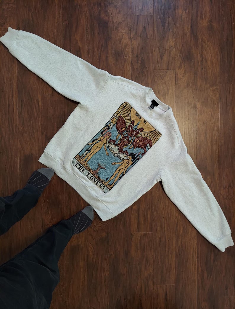 Puede incluir: Una sudadera gris claro con una gran imagen bordada de la carta del tarot de Los Enamorados. La carta est&aacute; en un estilo tradicional con un fondo azul y detalles dorados. El texto "THE LOVERS" est&aacute; impreso debajo de la carta.