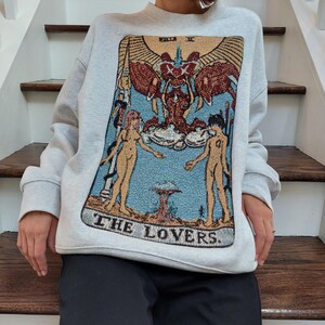 Puede incluir: Una sudadera gris con una gran imagen bordada de la carta del tarot de Los Enamorados. La carta presenta dos figuras desnudas de pie con los brazos en alto, una figura alada sobre ellas, y el texto "THE LOVERS."