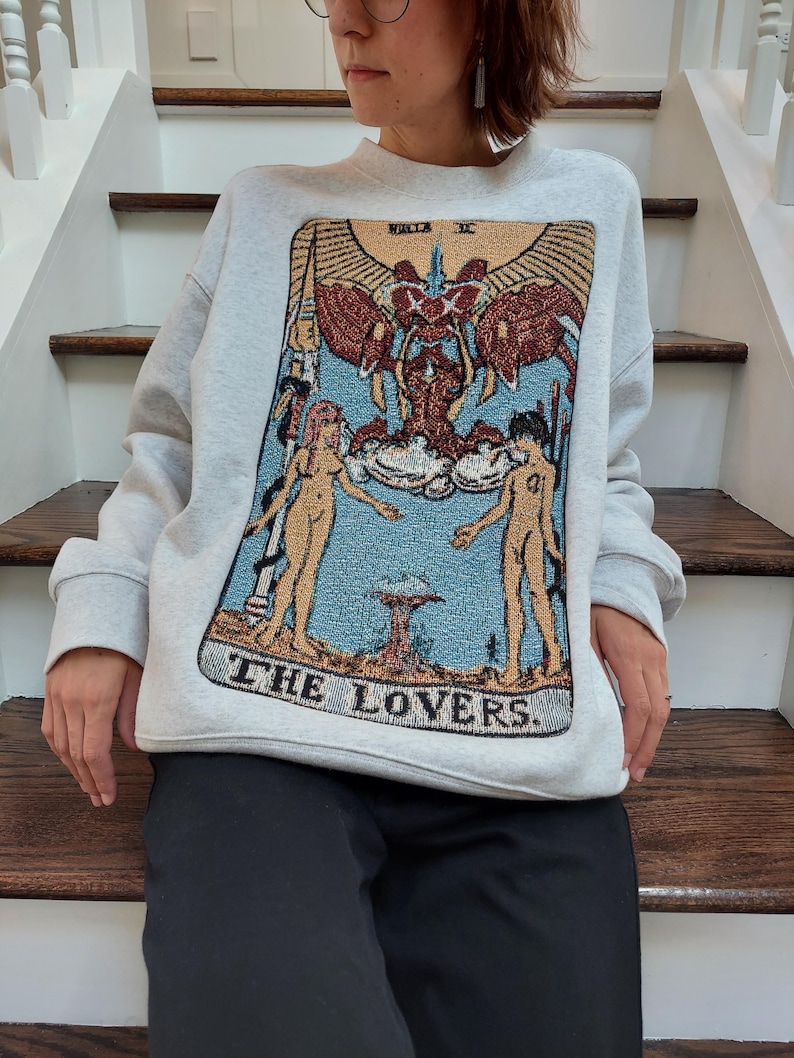 Puede incluir: Una sudadera gris con una gran imagen bordada de la carta del tarot de Los Enamorados. La carta presenta dos figuras desnudas de pie con los brazos en alto, una figura alada sobre ellas, y el texto "THE LOVERS."