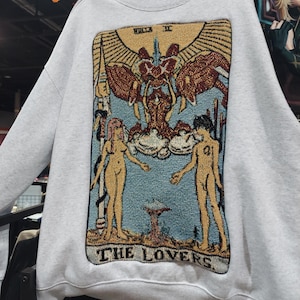 Puede incluir: Una sudadera blanca con una imagen de estilo tapiz de la carta del tarot de Los Enamorados. La carta presenta dos figuras desnudas de pie a cada lado de un &aacute;ngel alado con un halo. La carta est&aacute; rodeada de un marco dorado y el texto "THE LOVERS" est&aacute; impreso en la parte inferior.