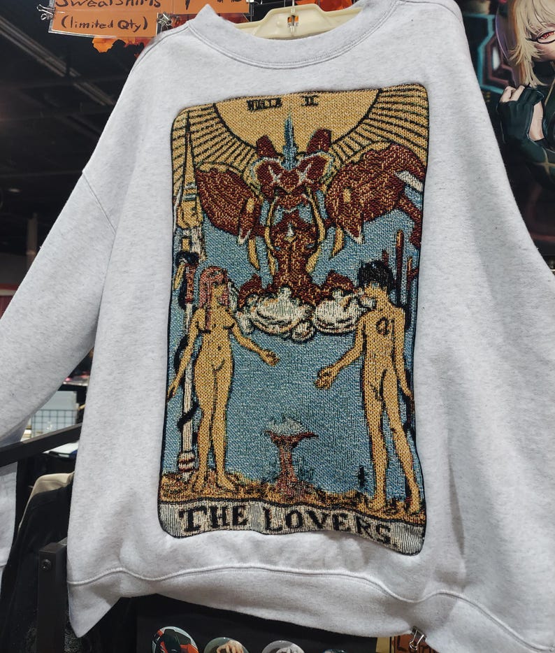 Puede incluir: Una sudadera blanca con una imagen de estilo tapiz de la carta del tarot de Los Enamorados. La carta presenta dos figuras desnudas de pie a cada lado de un &aacute;ngel alado con un halo. La carta est&aacute; rodeada de un marco dorado y el texto "THE LOVERS" est&aacute; impreso en la parte inferior.