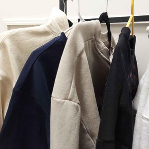 Puede incluir: Cinco sudaderas diferentes colgadas en un estante. Las sudaderas son de varios colores, incluyendo beige, azul marino, negro y blanco.