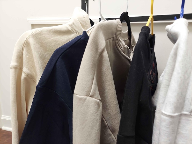 Puede incluir: Cinco sudaderas diferentes colgadas en un estante. Las sudaderas son de varios colores, incluyendo beige, azul marino, negro y blanco.