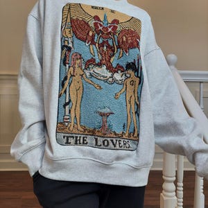 Puede incluir: Una sudadera gris con una imagen de estilo tapiz de la carta del tarot de Los Enamorados. La carta presenta dos figuras, un &aacute;ngel alado y el texto "THE LOVERS".