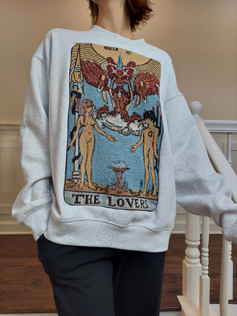 Puede incluir: Una sudadera gris con una imagen de estilo tapiz de la carta del tarot de Los Enamorados. La carta presenta dos figuras, un &aacute;ngel alado y el texto "THE LOVERS".