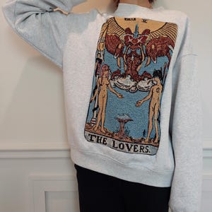 Puede incluir: Una sudadera gris con una gran imagen bordada de la carta del tarot de Los Enamorados. La carta presenta dos figuras de pie a cada lado de una figura alada con un halo. El texto "THE LOVERS" est&aacute; impreso debajo de la imagen.