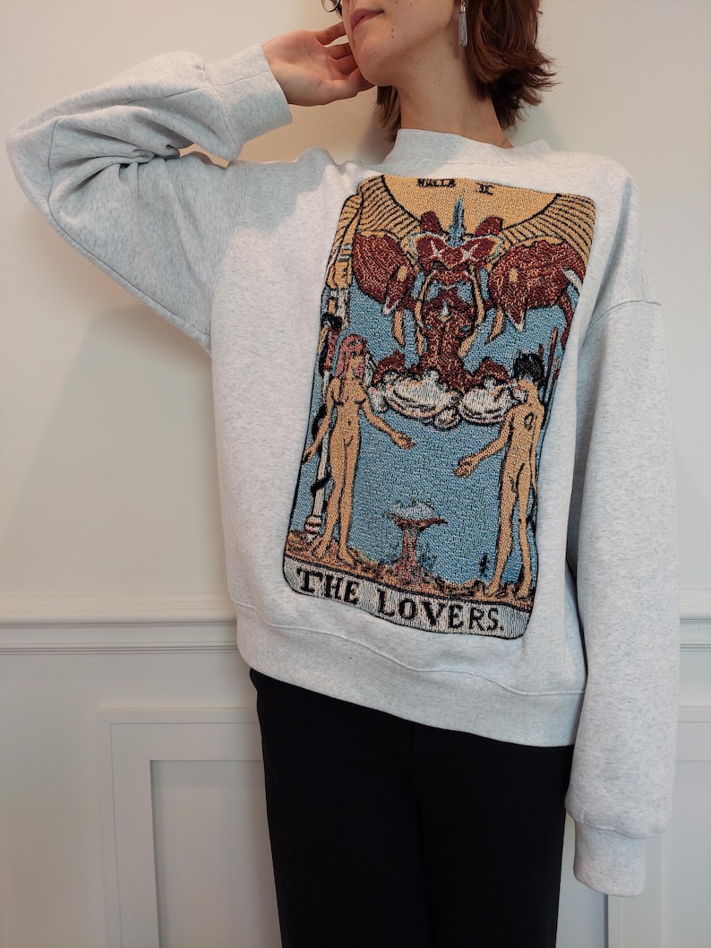 Puede incluir: Una sudadera gris con una gran imagen bordada de la carta del tarot de Los Enamorados. La carta presenta dos figuras de pie a cada lado de una figura alada con un halo. El texto "THE LOVERS" est&aacute; impreso debajo de la imagen.