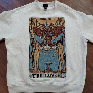 Puede incluir: Una sudadera gris con una imagen de estilo tapiz de la carta del tarot Los Amantes. La carta presenta dos figuras, un hombre y una mujer, de pie a cada lado de una figura alada. El texto "THE LOVERS" est&aacute; impreso debajo de la imagen.