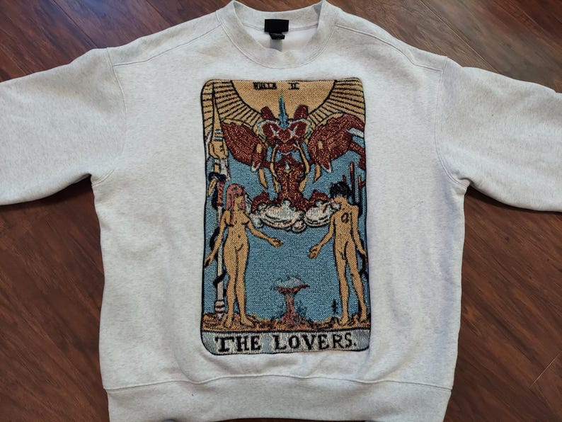 Puede incluir: Una sudadera gris con una imagen de estilo tapiz de la carta del tarot Los Amantes. La carta presenta dos figuras, un hombre y una mujer, de pie a cada lado de una figura alada. El texto "THE LOVERS" est&aacute; impreso debajo de la imagen.