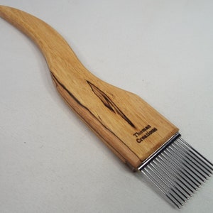 May include: A wooden fiber comb with a curved handle and a row of metal tines. The comb is made of light-colored wood with dark grain patterns. The text "Thomas Creations" is printed on the handle.