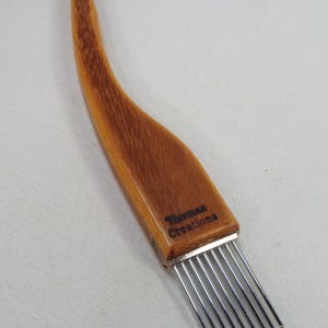 May include: A wooden scoring tool with a curved handle featuring the text Thomas Creations printed in black. The tool has a metal ferrule attached to a row of seven thin, parallel, silver-colored metal tines extending from the base of the handle.