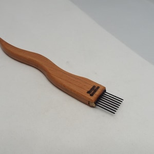 May include: A wooden scoring tool with a wavy, ergonomic handle and a row of six thin, black metal tines extending from one end. The wood features a natural grain pattern, and the brand name is stamped in black ink near the metal tines.