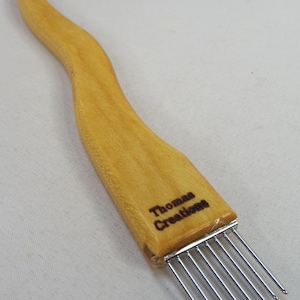 May include: A wooden weaving tool with a curved handle and a row of metal tines. The handle is light brown with the text "Thomas Creations" printed on it. The tines are silver and have small, rounded tips.