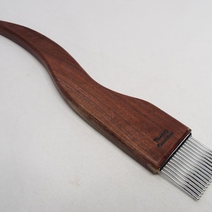May include: A wooden weaving comb with a curved handle and a row of metal teeth. The comb is made of dark brown wood and has the text "Thomas Gummert" printed on the handle.