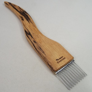 May include: A wooden weaving comb with a curved handle and metal tines. The comb is made of light-colored wood with dark grain patterns. The text "Thomas Creations" is visible on the handle.