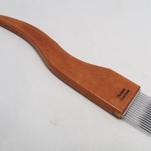 May include: A wooden weaving comb with a curved handle and a rectangular head with metal teeth. The comb is made of reddish-brown wood and has the text "Thomas Creations" on the head.