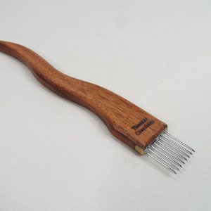 May include: A wooden weaving tool with a curved handle and a row of metal tines. The handle is a warm brown color, and the text "Theitti Creations" is visible on the handle. The tool is designed for fiber art and weaving projects.