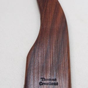 May include: A wooden weaving comb with a curved handle and a row of metal teeth. The comb is a dark brown color and has the text "Thomas Creations" engraved on the handle.