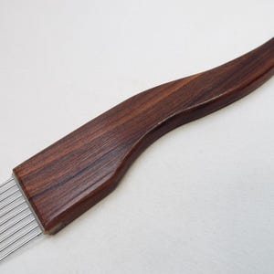 May include: A wooden weaving comb with a curved handle and a rectangular head with evenly spaced metal tines. The comb is made of dark brown wood with a visible grain pattern, and the tines are silver.
