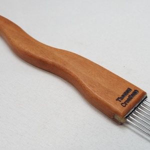 May include: A wooden weaving tool with a curved handle and a row of metal tines. The tool is a light brown color and has the text "Thomas Creations" printed on the side. The tool is designed for fiber art.