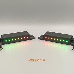 Könnte beinhalten: Zwei schwarze, rechteckige elektronische Sensoreinheiten mit Befestigungslaschen, jede mit einer Reihe von acht leuchtenden LED-Lichtern in Grün, Gelb und Rot. Jede Einheit hat ein schwarzes Stromkabel und zwei kleine Metallschrauben. Version A.