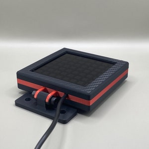 K&ouml;nnte beinhalten: Ein quadratisches Ger&auml;t in Schwarz und Rot mit einem schwarzen gitterartigen Display und einem schwarzen Kabel, das an der Seite befestigt ist.