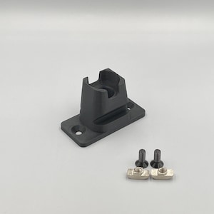 Könnte beinhalten: Eine schwarze Wandhalterung aus Kunststoff mit zwei Schrauben und zwei Muttern aus Metall. Die Halterung hat eine rechteckige Basis und einen erhöhten Abschnitt mit einem Loch in der Mitte.