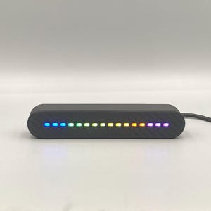 Peut inclure: Un appareil électronique rectangulaire noir aux extrémités arrondies. Il présente une série de petites lumières espacées qui affichent un dégradé de couleurs, du bleu au violet. Un câble noir s'étend de l'appareil.