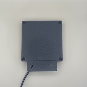 K&ouml;nnte beinhalten: Ein schwarzes rechteckiges Ger&auml;t mit einem Karbonfaser-Muster und Schraubenl&ouml;chern an den Seiten. Das Ger&auml;t hat ein Kabel.