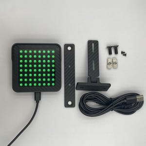 May include: A square black device with a carbon fiber texture and a grid of sixty-four illuminated green LED lights. Included are a black mounting bracket, a T-shaped stand, two silver T-nuts, four black screws, and a black USB power cable.