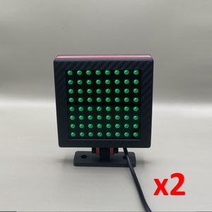 Puede incluir: Un panel de luz LED negro y rojo con una cuadrícula de luces verdes. El panel está montado en un soporte negro y tiene un cable negro. El texto "x2" está escrito en rojo debajo del panel.
