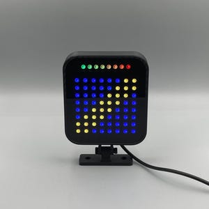 Puede incluir: Una luz LED cuadrada negra con una cuadrícula de luces azules, amarillas y verdes. La luz está montada en un soporte negro y tiene un cable negro.