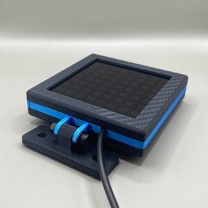 K&ouml;nnte beinhalten: Ein schwarzes quadratisches Ger&auml;t mit blauer Einfassung und einem schwarz-wei&szlig;en Gittermuster auf dem Display. Das Ger&auml;t ist an einer schwarzen Halterung befestigt und hat ein schwarzes Kabel.