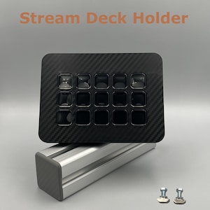 Könnte beinhalten: Ein schwarzer Stream Deck Halter mit Karbonfasermuster und 15 quadratischen Tasten. Der Halter steht auf einer silbernen Metallbasis, darunter sind zwei kleine silberne Schrauben zu sehen. Der Text "Stream Deck Holder" ist orange dargestellt.