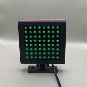 K&ouml;nnte beinhalten: Ein schwarzes quadratisches LED-Leuchtpanel mit einer Carbonfaser-Textur und einem roten Streifen. Das Panel hat ein Raster aus gr&uuml;nen LED-Leuchten.