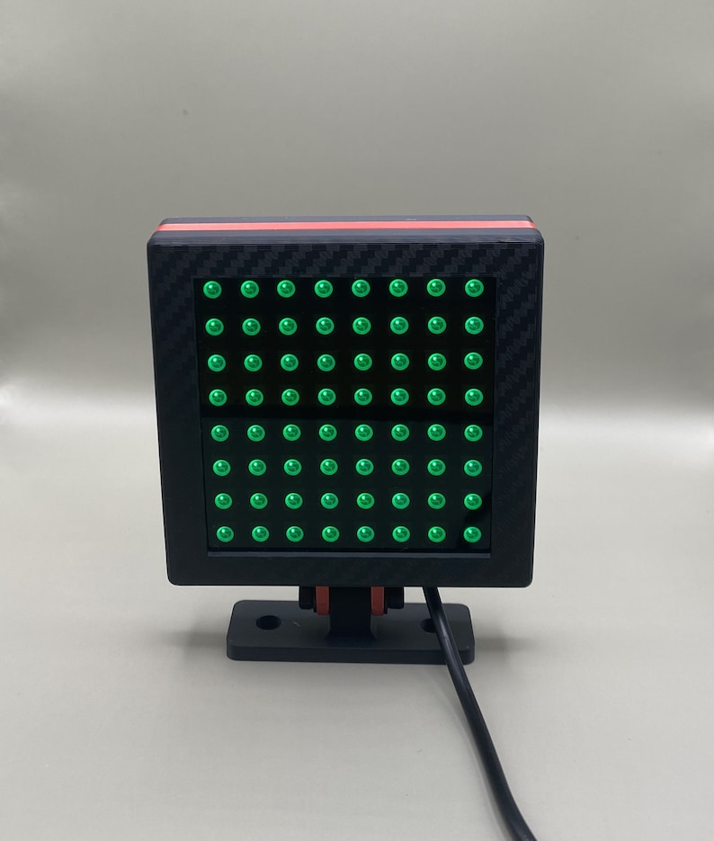 K&ouml;nnte beinhalten: Ein schwarzes quadratisches LED-Leuchtpanel mit einer Carbonfaser-Textur und einem roten Streifen. Das Panel hat ein Raster aus gr&uuml;nen LED-Leuchten.
