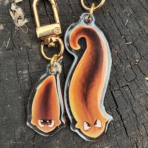 May include: Two clear acrylic keychains with gold-coloured hardware. Each keychain features a cartoonish, brown and orange creature design with white accents. One keychain is a teardrop shape, and the other is a larger, curved shape.
