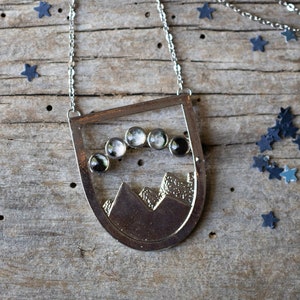 May include: A silver necklace with a pendant featuring a mountain range and a crescent moon with eight phases. The pendant is surrounded by small blue stars.