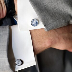 May include: A pair of cufflinks featuring a moon design. The cufflinks are silver with a blue and white image of the moon.