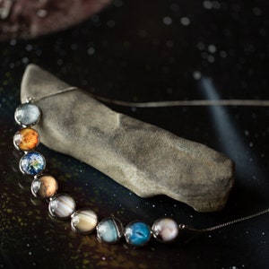 May include: A silver chain necklace with round glass beads featuring images of the planets in our solar system. The necklace is displayed on a gray rock against a dark background with white speckles.
