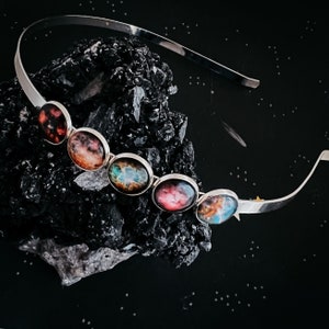 May include: A silver headband with six colorful galaxy-themed glass cabochons. The cabochons are arranged in a row and are set in silver bezels. The headband is on a black background.