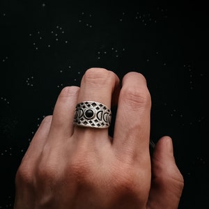 Könnte beinhalten: Ein silberner Ring mit einem schwarzen Stein in der Mitte und einem Design aus Mondsicheln und Sternen darum herum.