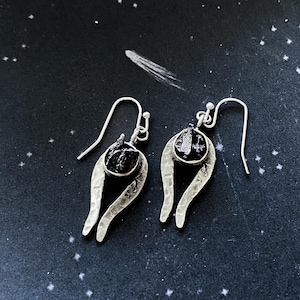 May include: A pair of silver earrings featuring black teardrop-shaped stones. The earrings have a distinctive, curved design.